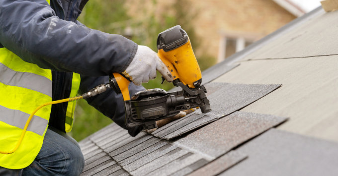 person in reflective vest, with gloves on, holding nail gun and working on shingling a roof