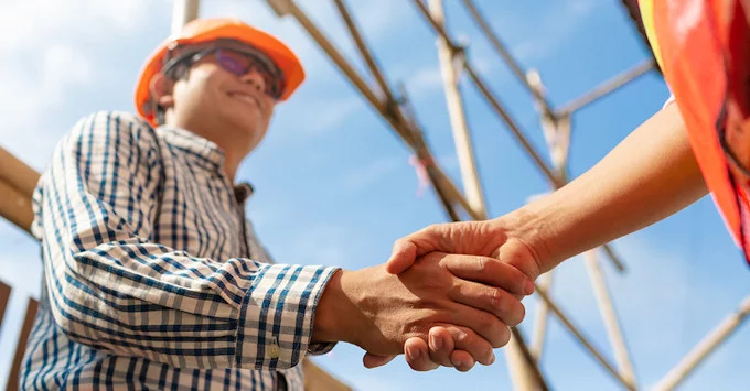 two people in construction zone shaking hands, one is visible with orange hard hat, protective glasses and a plaid shirt opposite person dressed in orange shirt